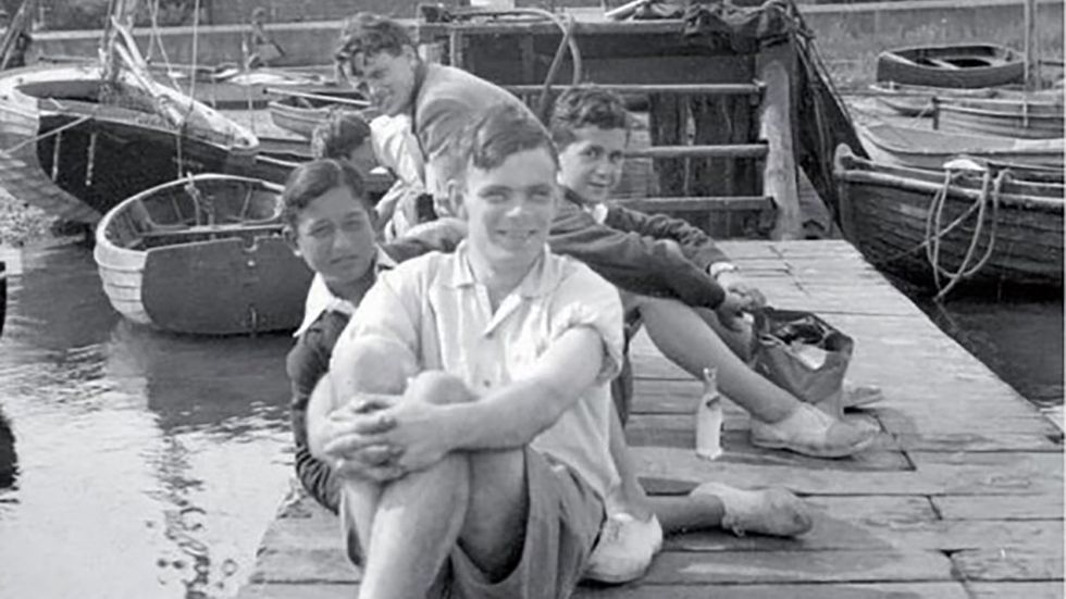 A young smiling Alan Turing and friends wearing shorts and boating shoes on a small dock surrounded by small boats